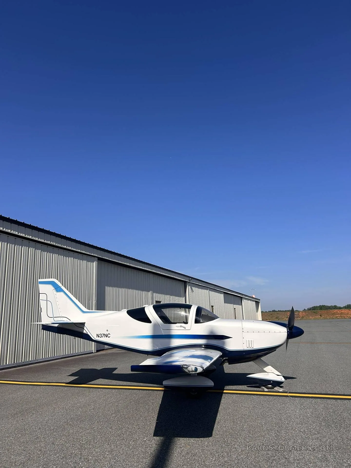 A white and blue single-engine aircraft with registration N37NC is parked on an asphalt tarmac next to a hangar, viewed from the side.