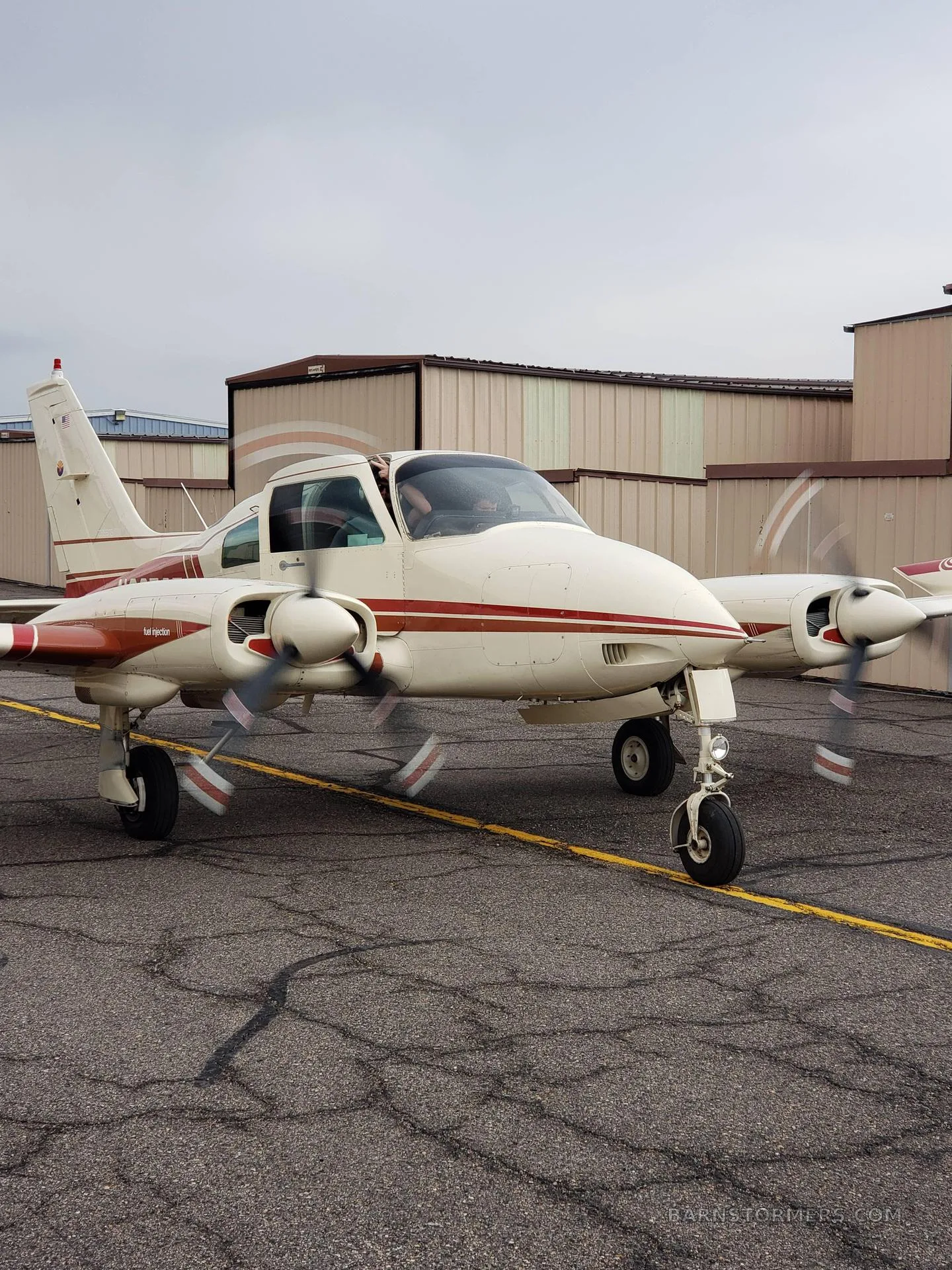 A white and red twin-engine propeller aircraft with spinning propellers is taxiing on an airport tarmac under a cloudy sky.