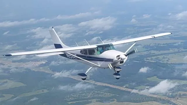 A white and blue Cessna 182 aircraft flies over a cloudy, green landscape.