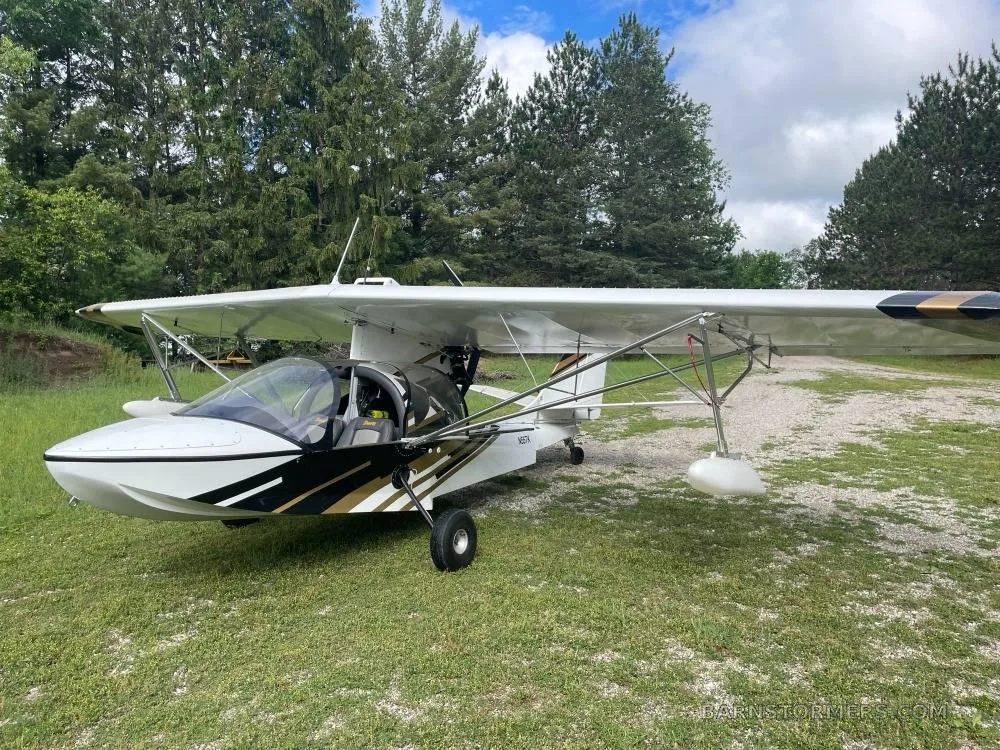 A white and gold amphibious light sport aircraft with floats and wheels parked on a grassy field with trees in the background.