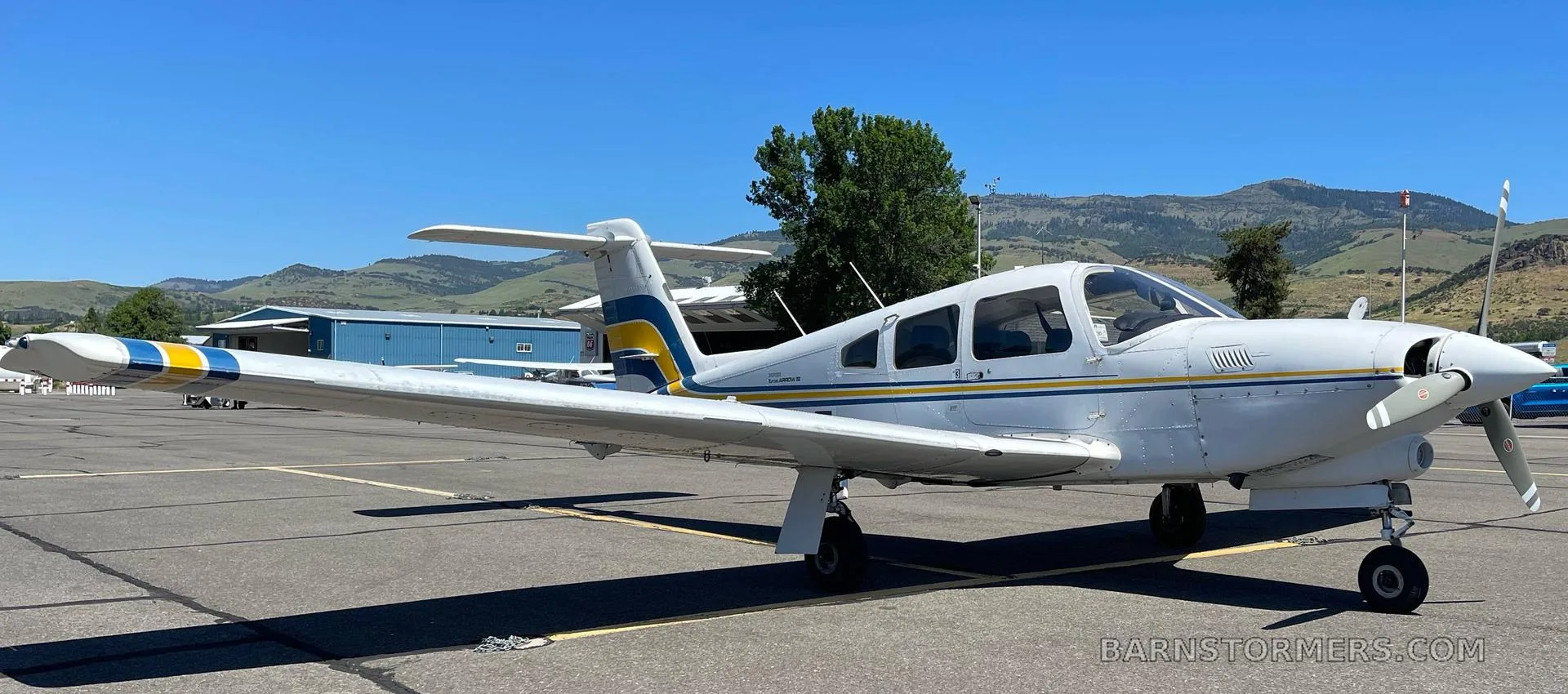 A white Piper Turbo Arrow III aircraft with blue and yellow stripes, registration N79AF, viewed from the front-right on an airfield.