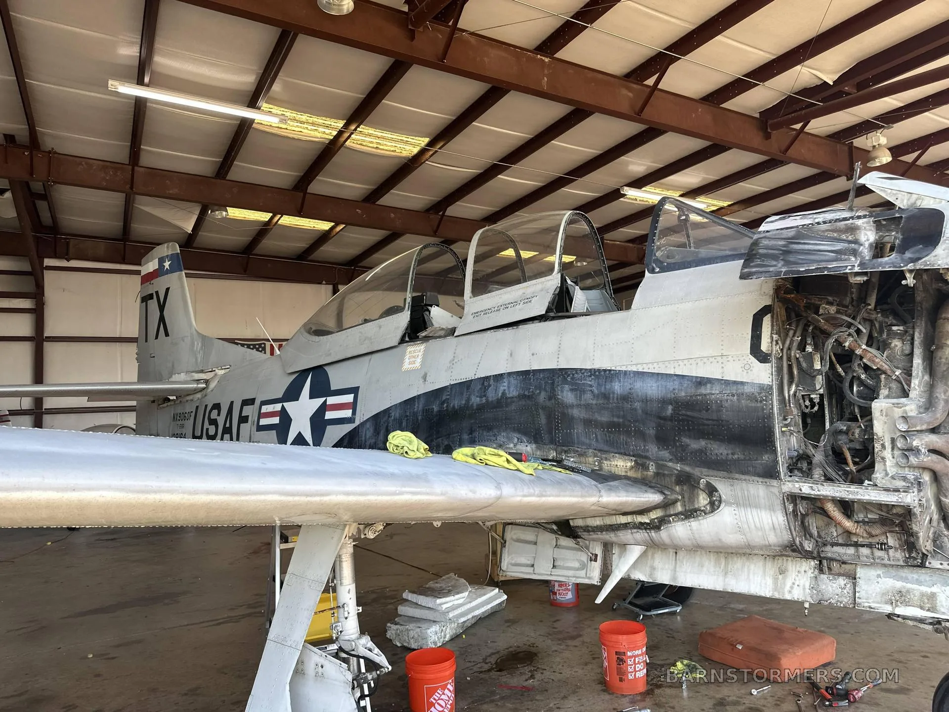 A gray military trainer aircraft, possibly a T-28 Trojan, with its engine cowling removed, sits in a hangar.