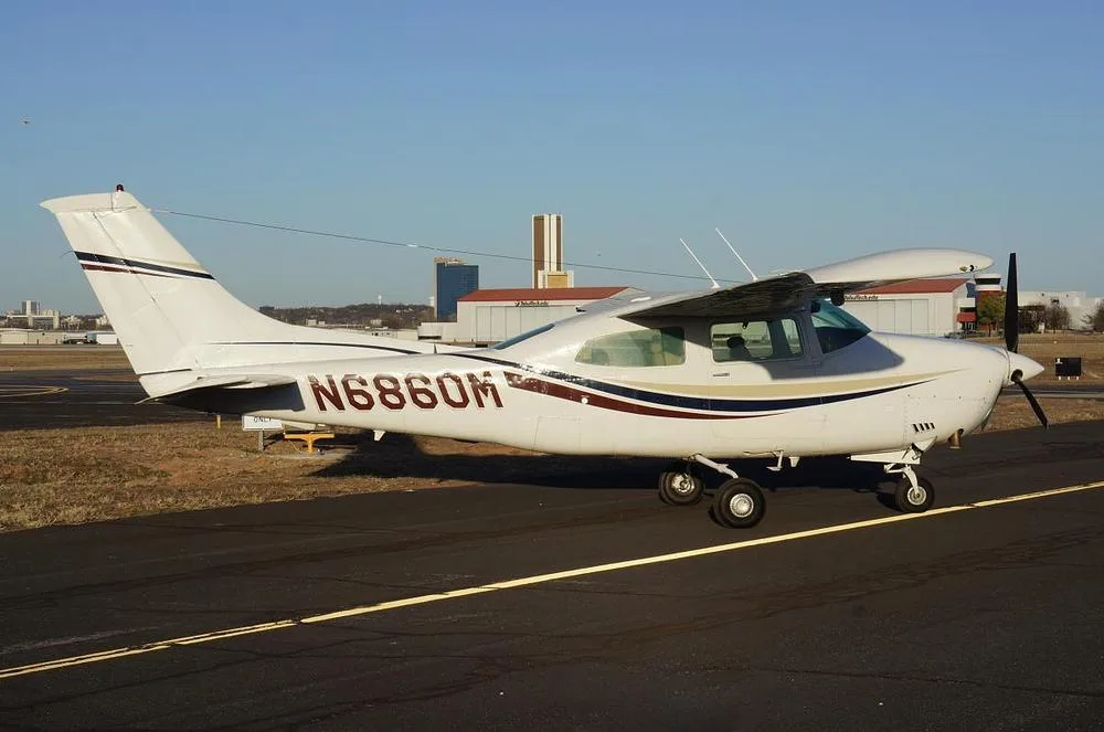 A white Cessna 210 Centurion aircraft with red, blue, and gold stripes on an airfield with distant buildings.