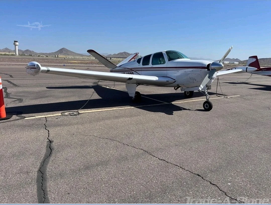 A front-quarter view of a white Beechcraft Bonanza with red stripes is parked on a tarmac with a control tower and mountains in the background.