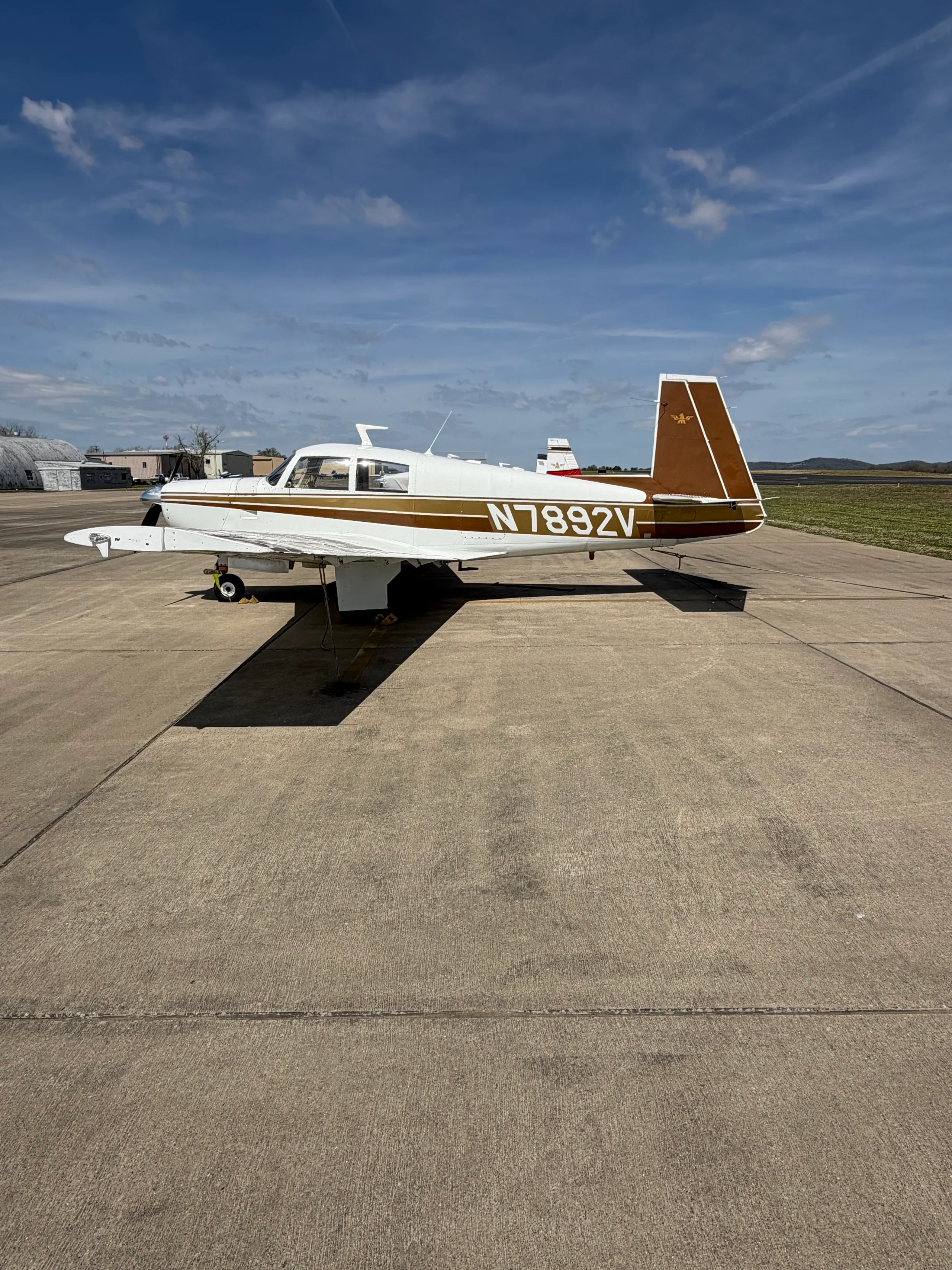 A white and brown single-engine aircraft (N7892V) parked on an asphalt tarmac under a partly cloudy sky.