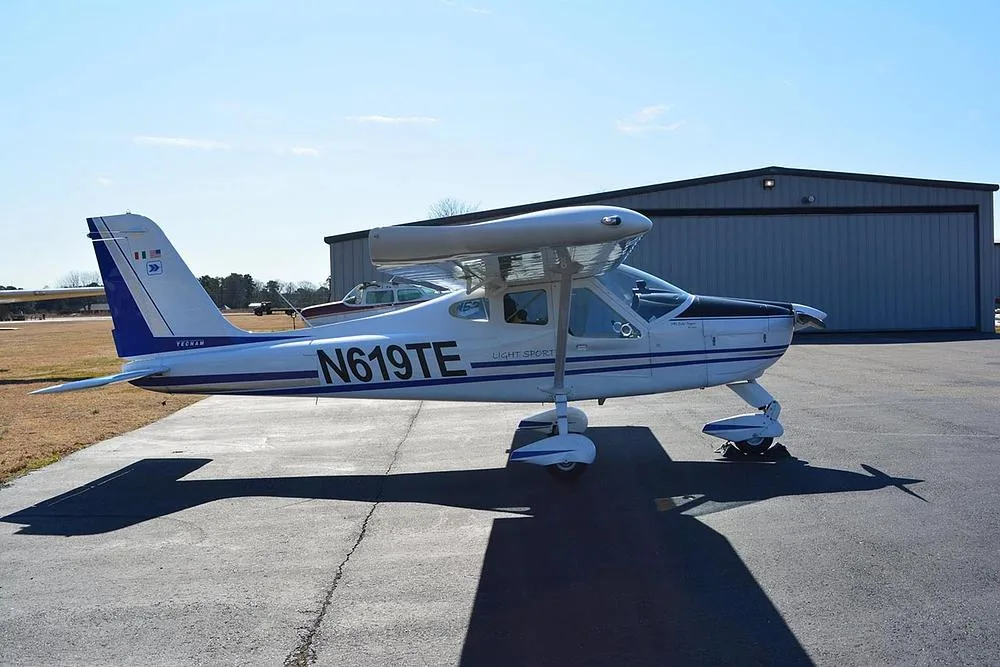 A white Tecnam Light Sport aircraft with blue and grey stripes parked on an asphalt ramp in front of a hangar.