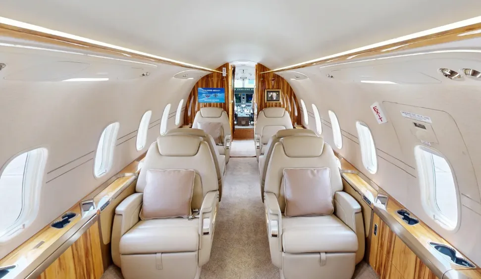 The spacious interior of a private jet cabin with two rows of light leather seats and wood accents.