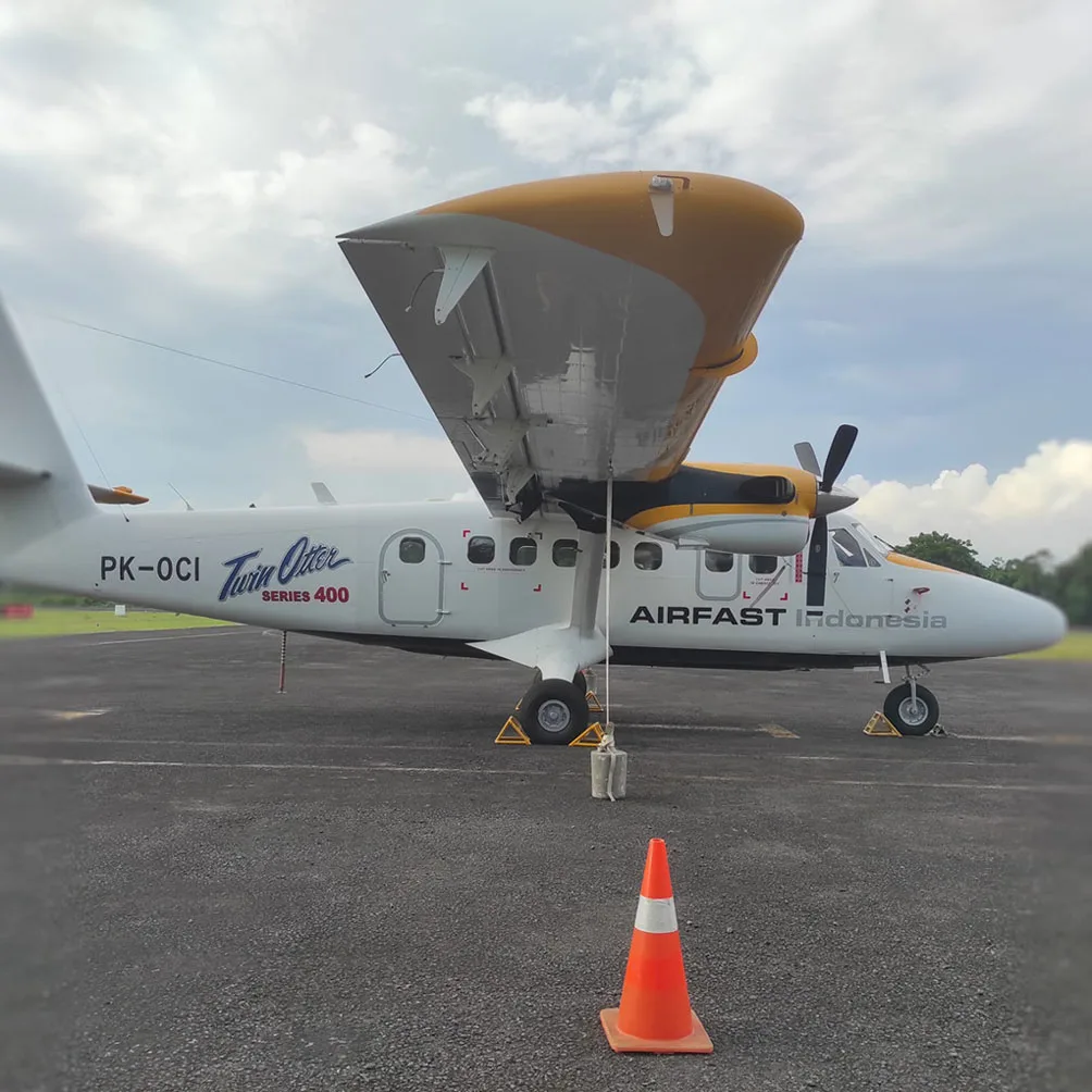 A side view of a white and yellow Airfast Indonesia Twin Otter Series 400 aircraft (PK-OCI) with its wing flaps extended, parked on an airfield next to an orange cone.