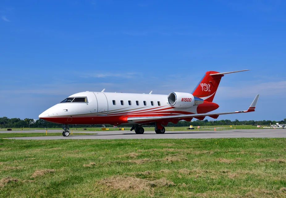 A white and red Bombardier Challenger business jet with gold accent stripes on a taxiway under a clear blue sky.