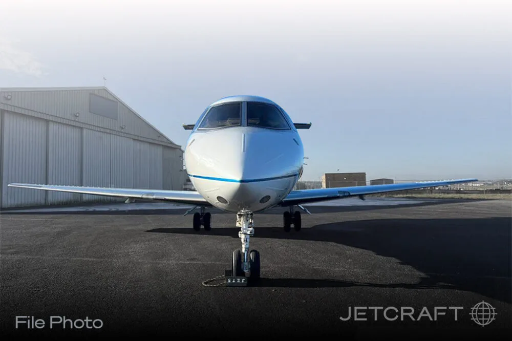 A white business jet with a blue stripe on the nose, parked on an asphalt tarmac in front of a hangar.