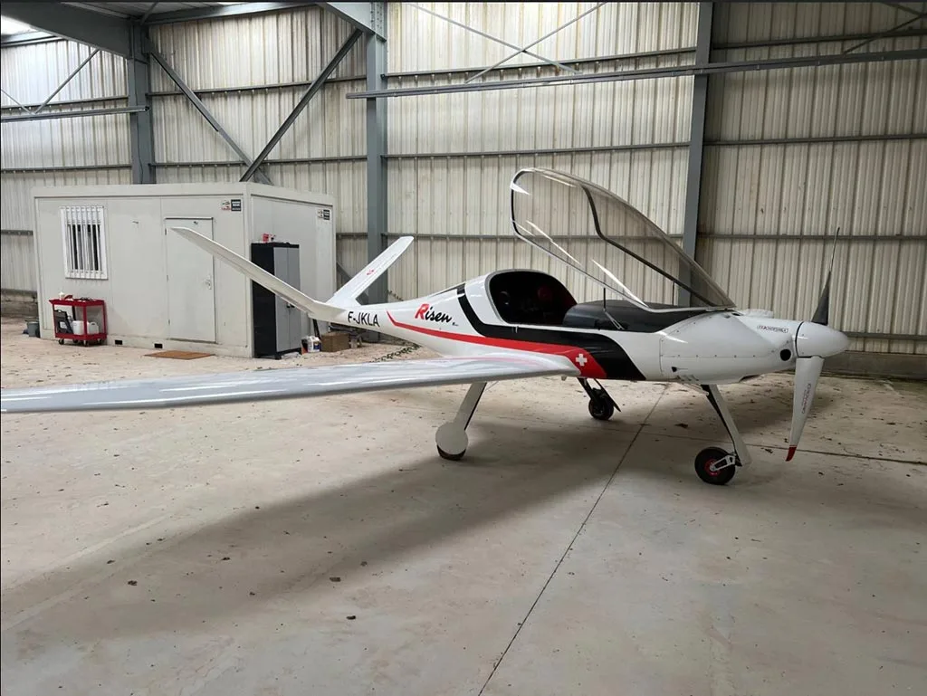 A white Risen aircraft with registration F-JKLA and an open canopy is parked inside a metal hangar.