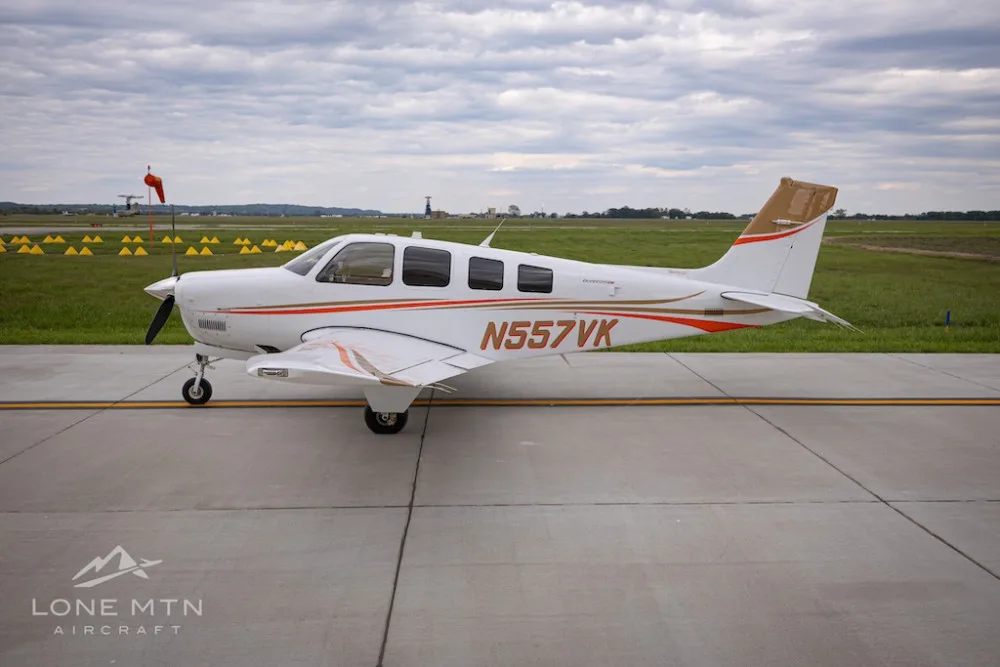 A side view of a white Beechcraft Bonanza G36 with orange and brown stripes, registration N557VK, parked on an airfield.
