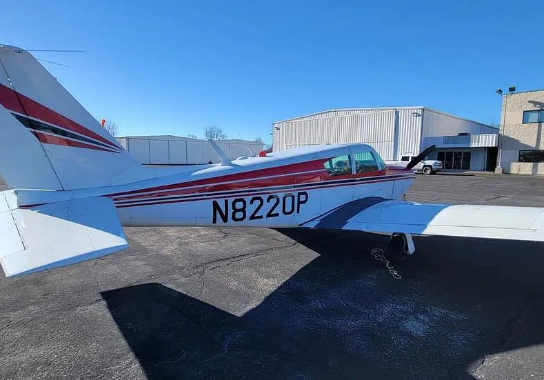 A rear-right view of a red and white Piper Comanche 250 aircraft, tail number N8220P, parked near a hangar.