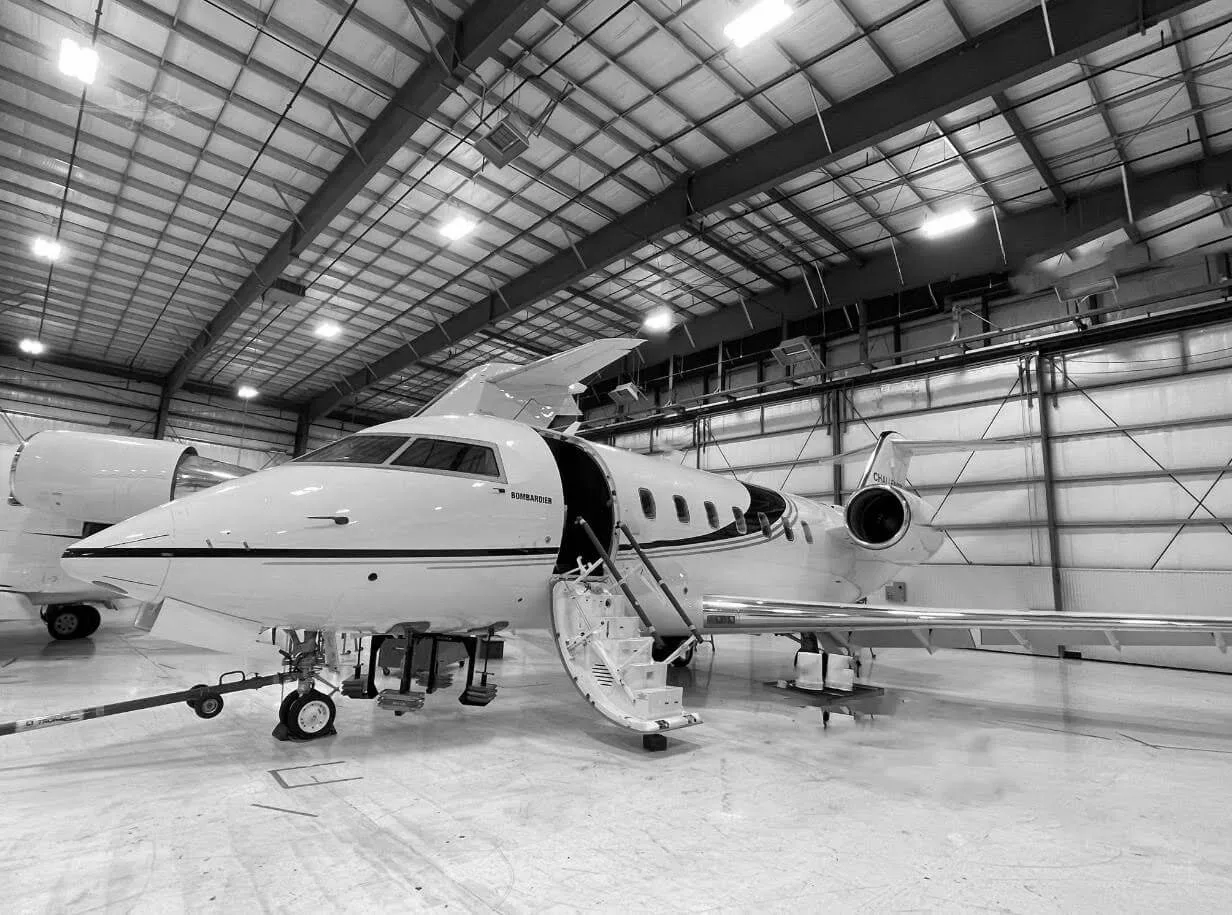 A black and white photo of a Bombardier business jet with its door open and stairs deployed, parked inside a hangar.