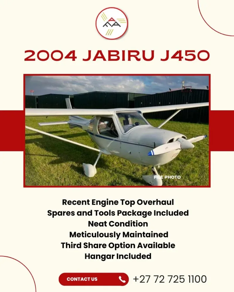 A white Jabiru J450 light aircraft with blue propeller tips is parked on a grassy field in front of green hangars.