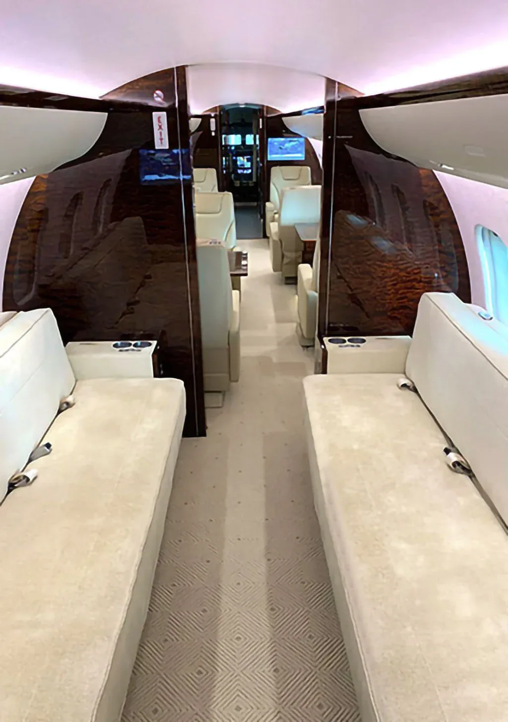A view down the aisle of a private jet cabin, showing cream divans and individual seats with dark wood accents.