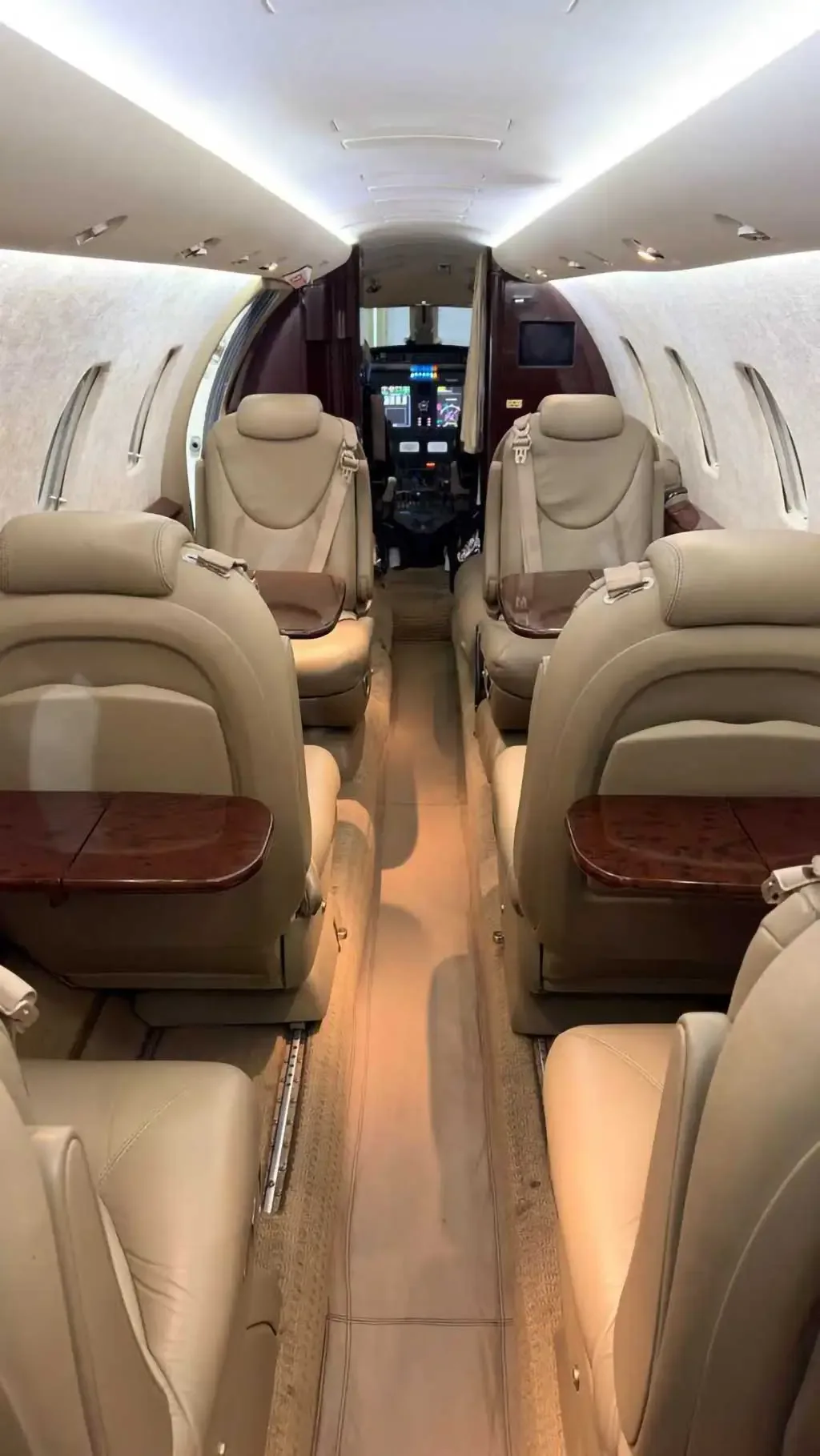 View of the aircraft cabin interior with beige leather seats and wooden tables, looking towards the cockpit.