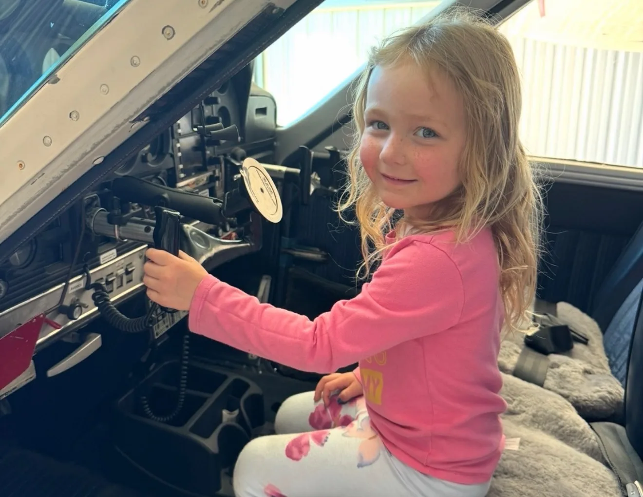 A young girl with blonde hair sits in the pilot's seat of an aircraft cockpit, smiling at the camera.