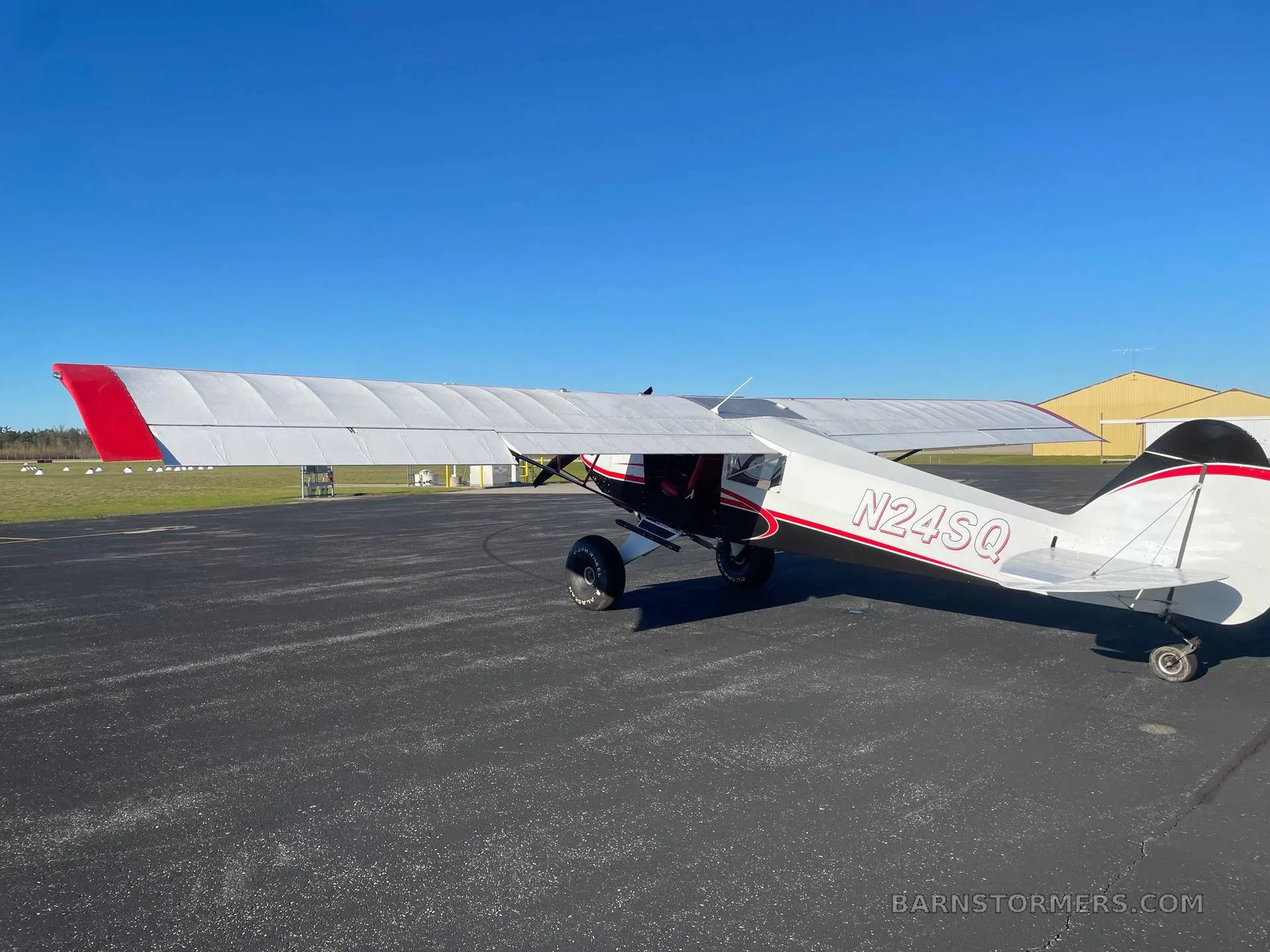A rear-side view of a white and red high-wing aircraft with registration N24SQ, parked on an asphalt apron with a distant hangar.
