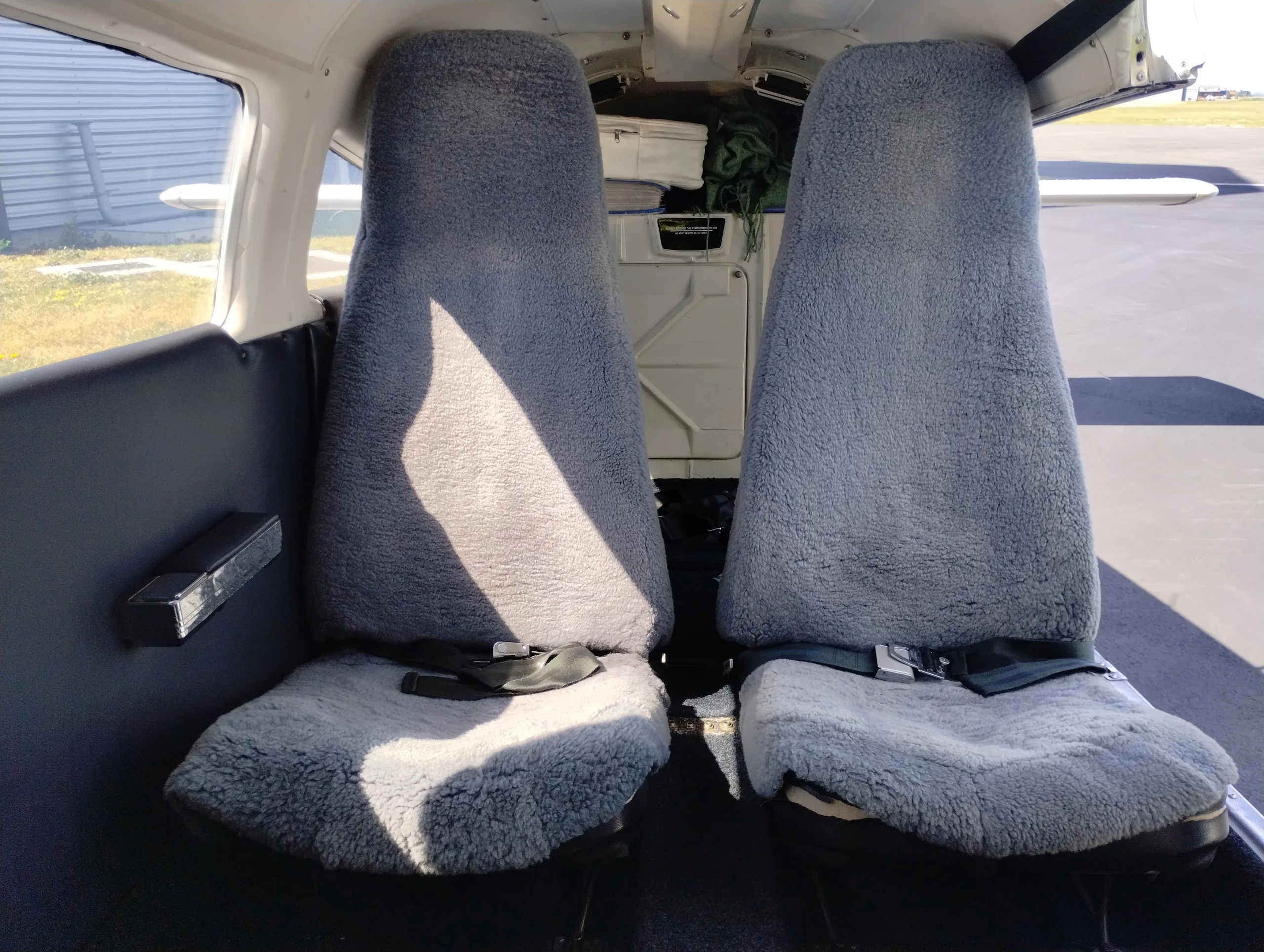 Two grey sheepskin-covered aircraft seats in a small plane's cabin, with a view of the exterior through the window.