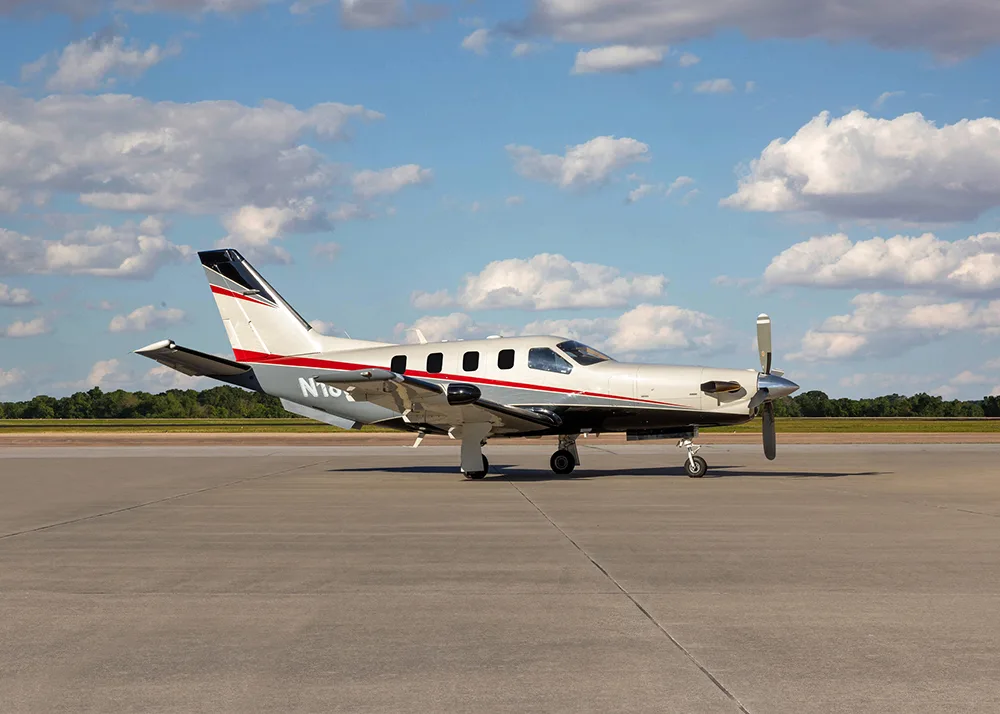 A white and grey TBM 900 series aircraft with red stripes, registration N180MF, parked on an airfield, viewed from the front-left.