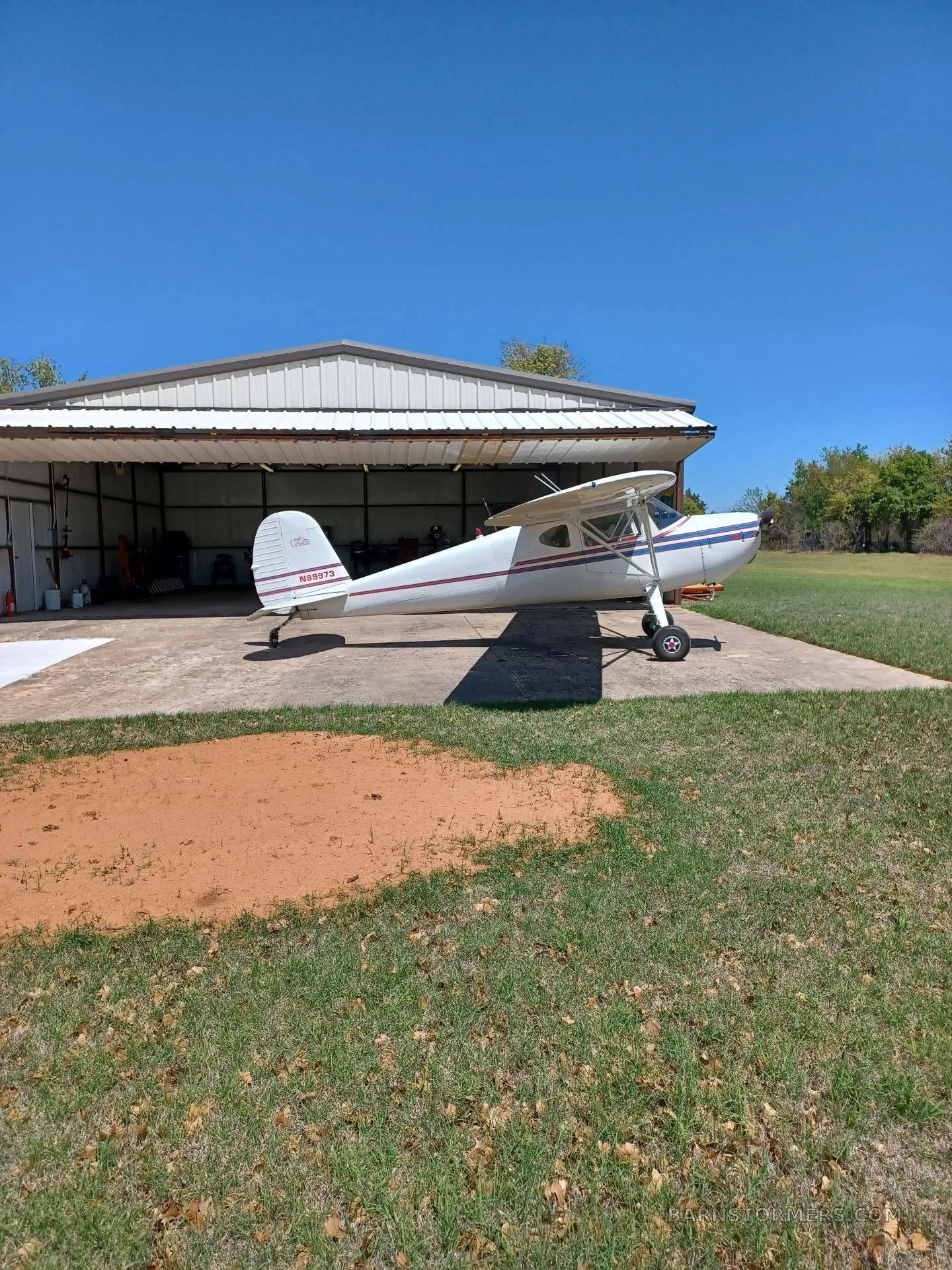 A white high-wing taildragger aircraft with blue and red stripes, tail number N6297D, parked on a concrete pad outside an open hangar.