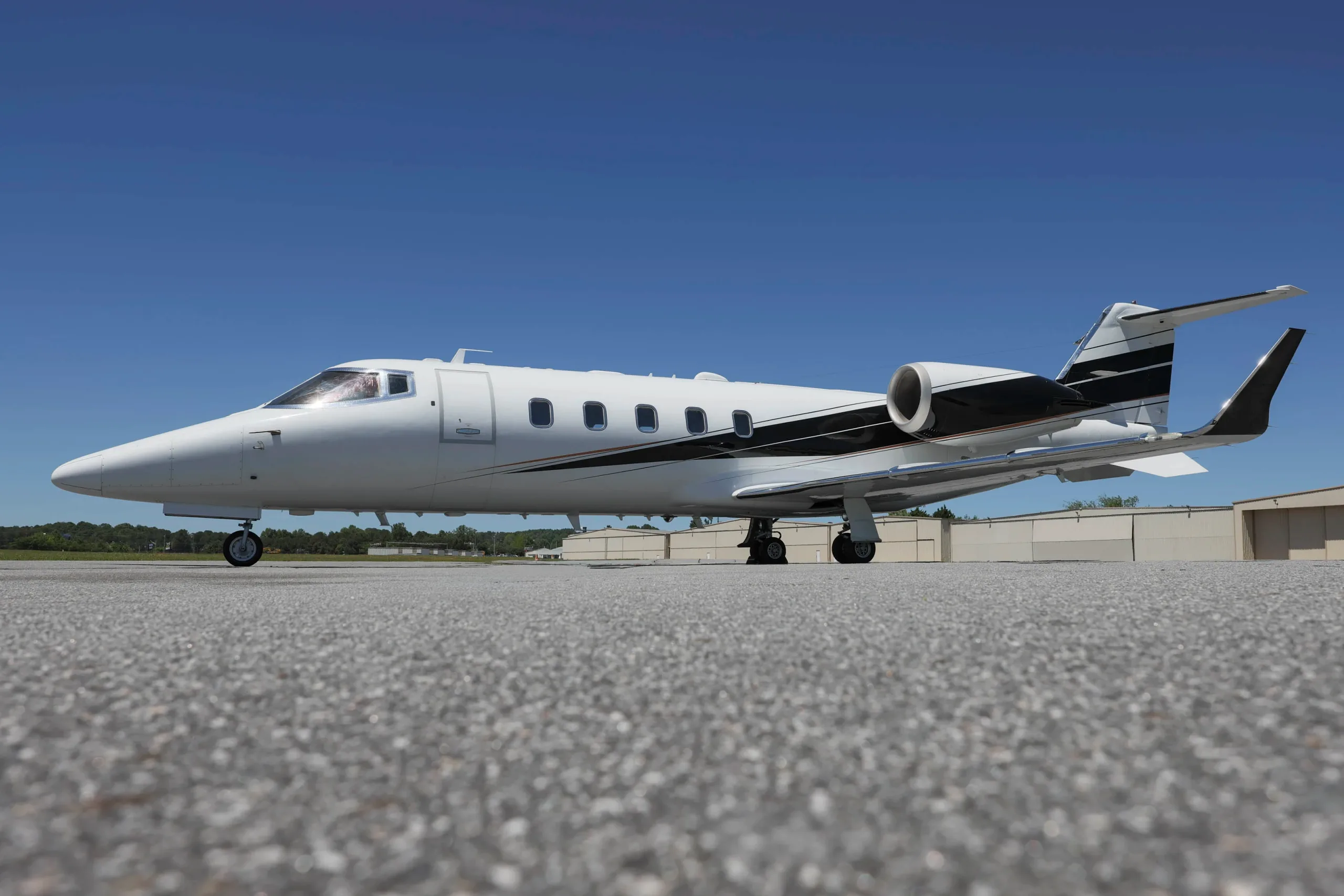 A white business jet with black and gold stripes on the side, parked on an asphalt tarmac under a clear blue sky.