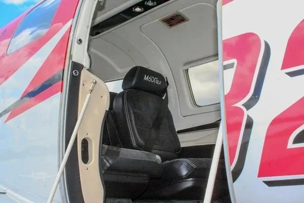 The open cabin door of a red and white Piper M600 SLS aircraft reveals a black leather passenger seat with "M600SLS" embroidered on the headrest.