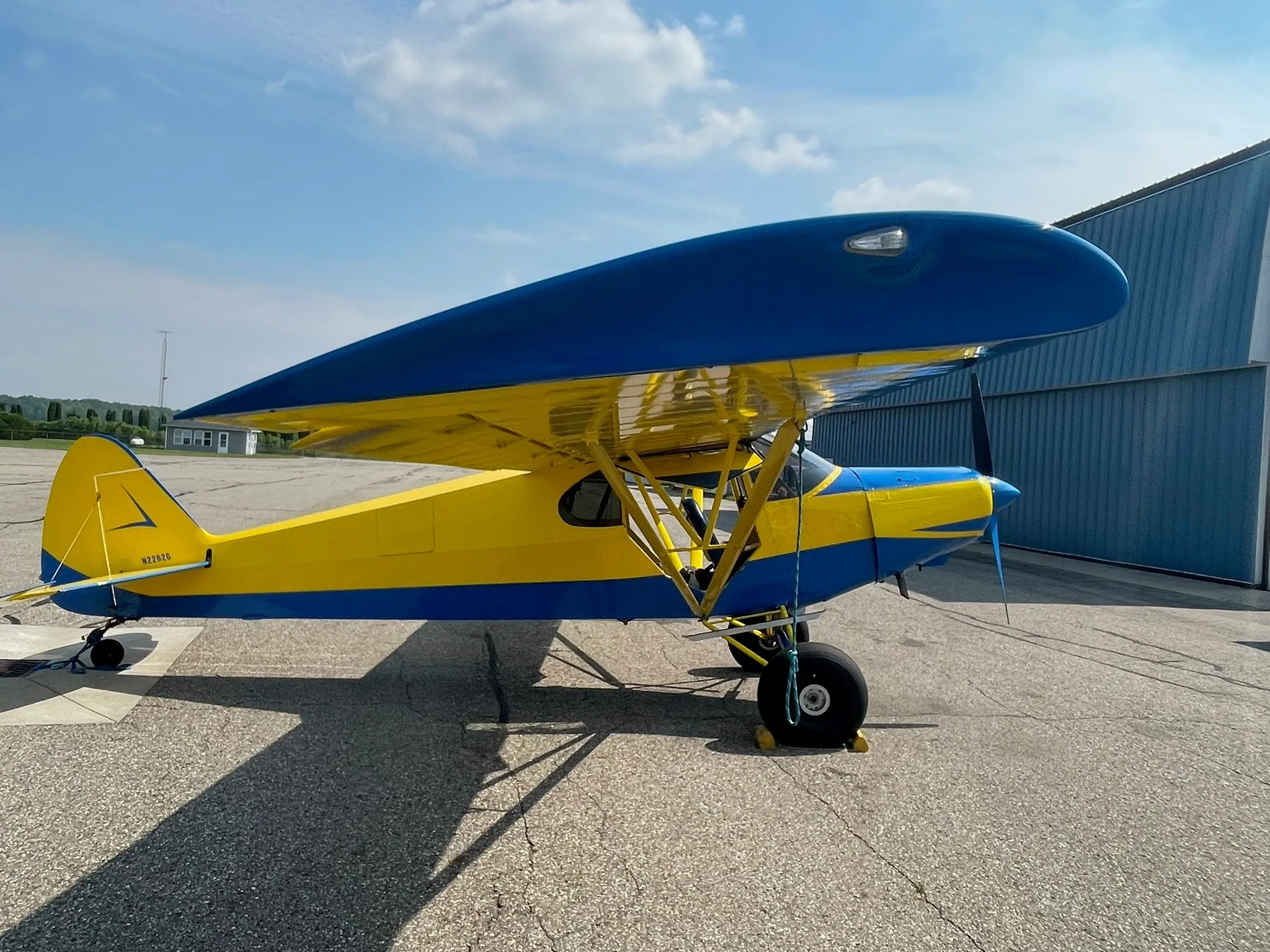 A yellow and blue Piper Super Cub (N2262G) parked on an airfield tarmac with a hangar in the background.