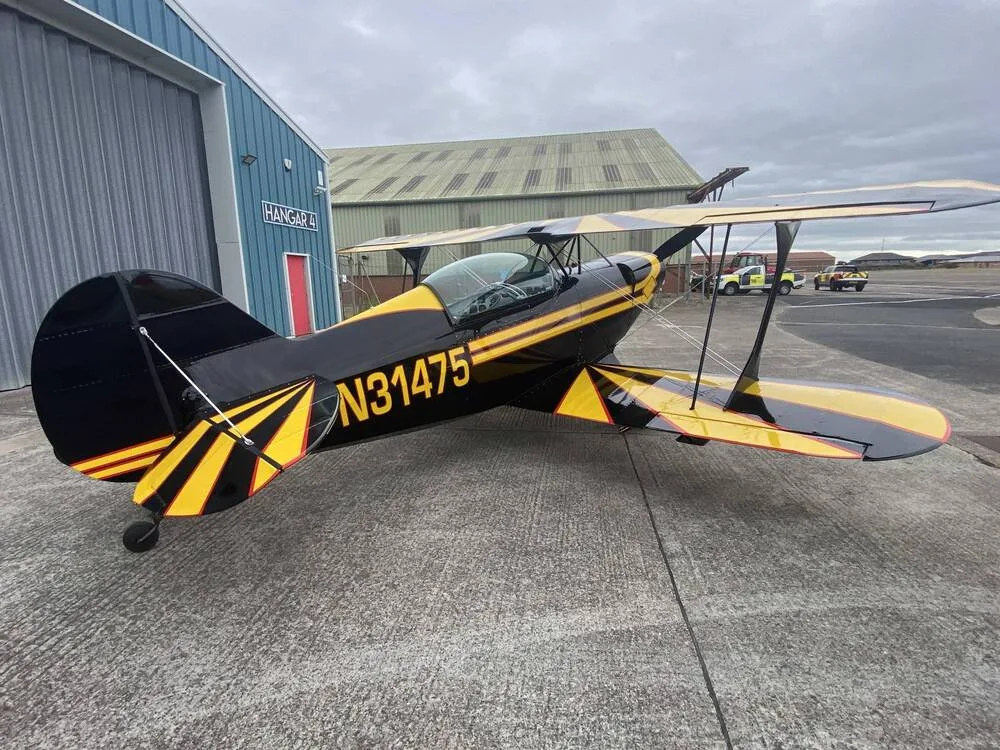 A black and yellow biplane, N31475, is parked on an airfield with a hangar in the background.