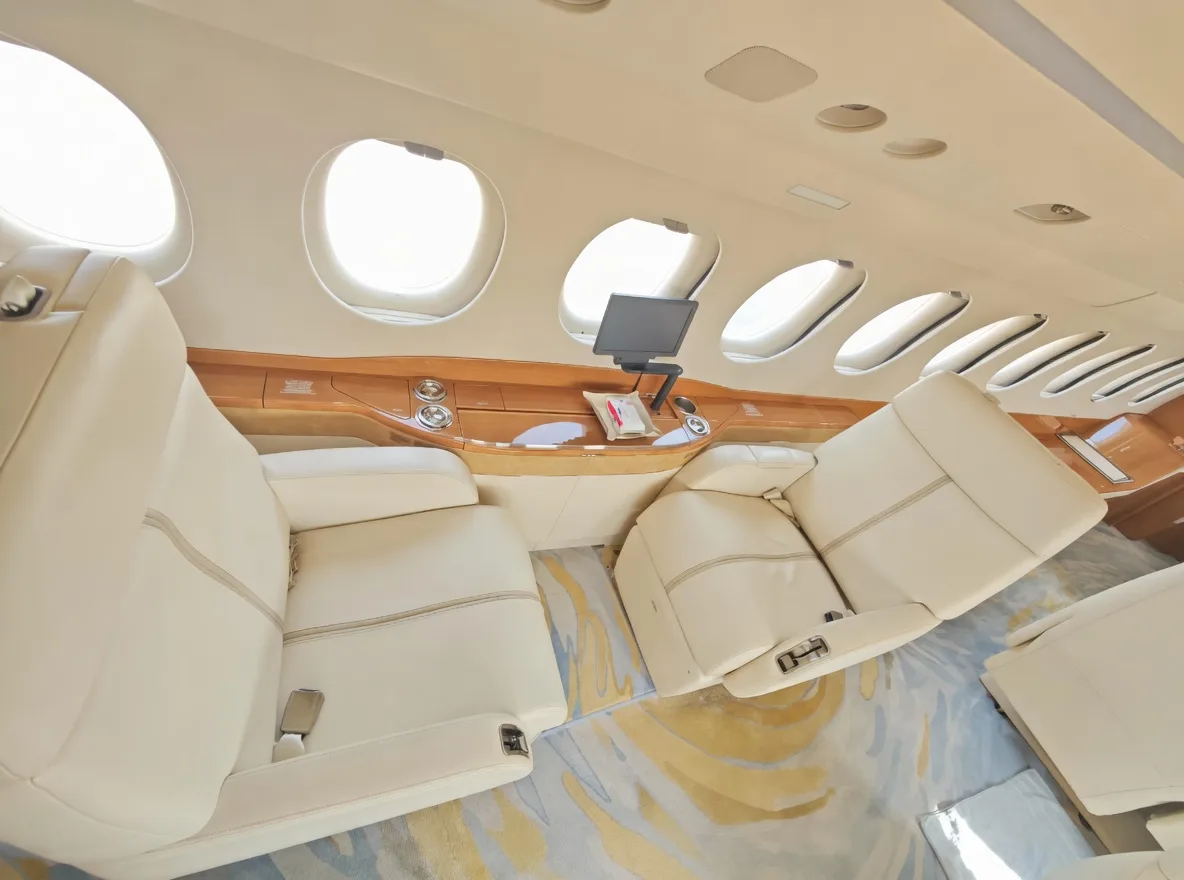A view inside an aircraft cabin showing cream leather seats, a wooden side panel, and a mounted display screen.