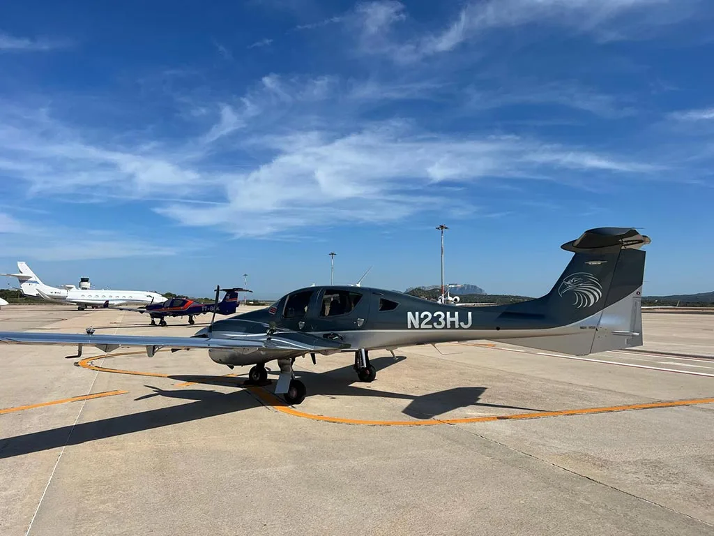 A side view of a dark grey Diamond DA62 aircraft with registration N23HJ and an eagle graphic on the tail, parked on an airport tarmac.