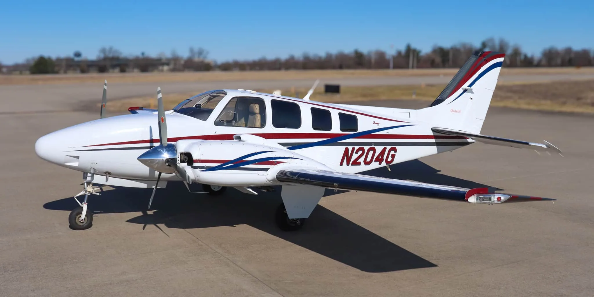 A white twin-engine Beechcraft Baron N204G with red, blue, and gray stripes, parked on an asphalt runway.