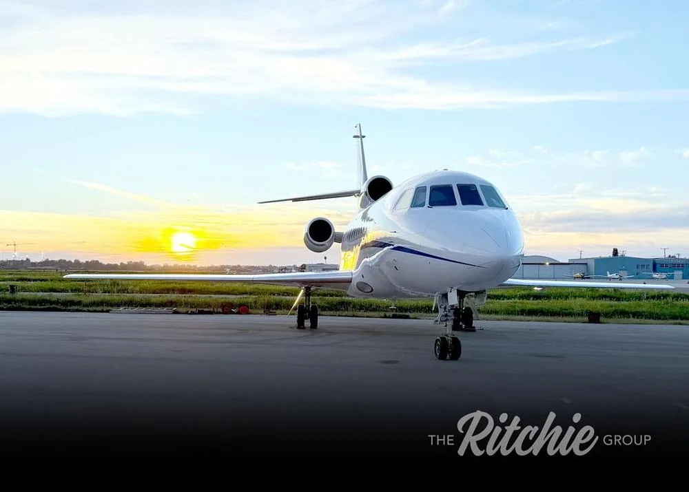A white Dassault Falcon business jet with blue accents seen from the front on a tarmac at sunset.