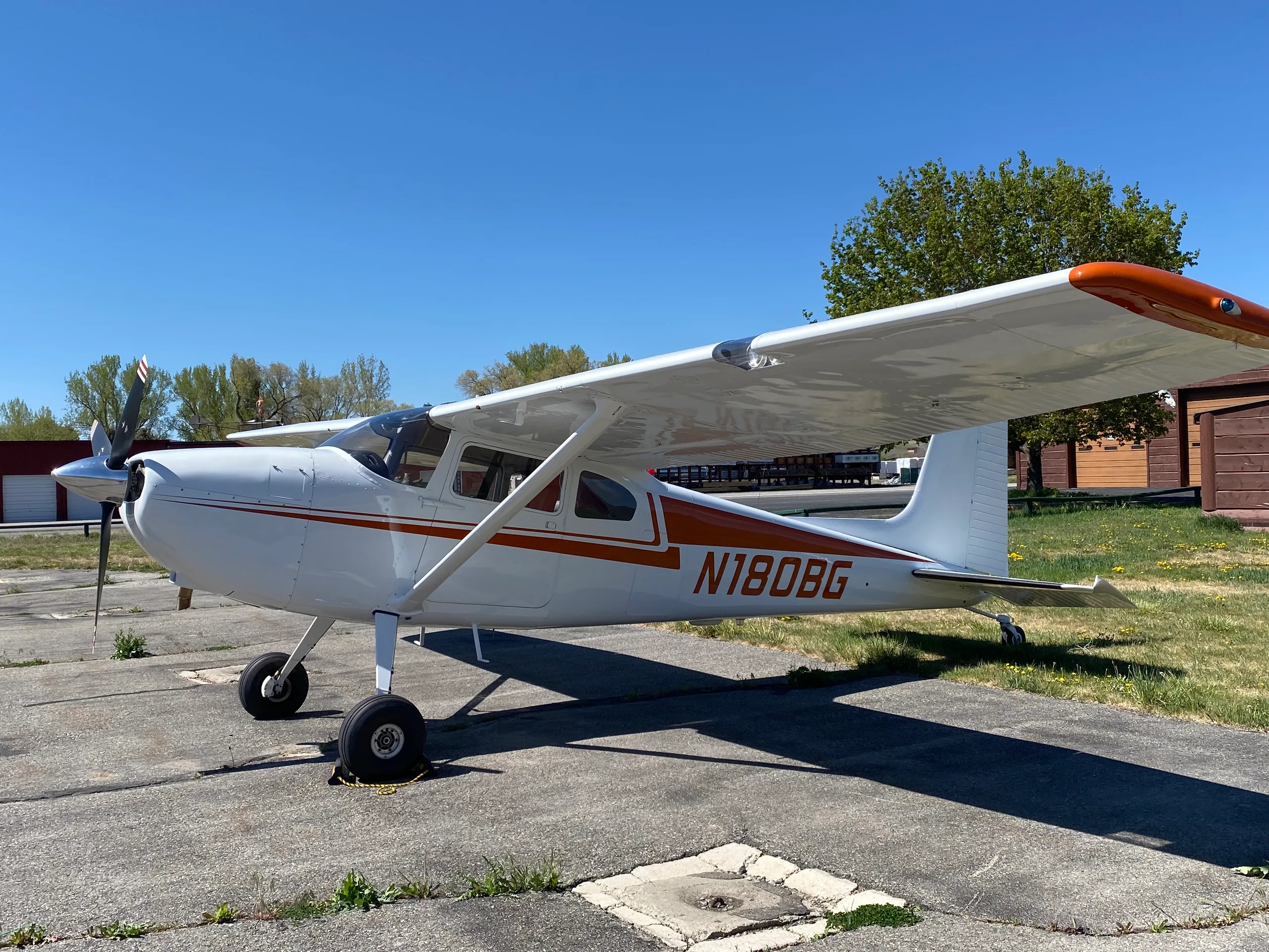 A white Cessna 170B with orange stripes and N180BG registration parked on an airfield under a clear blue sky.