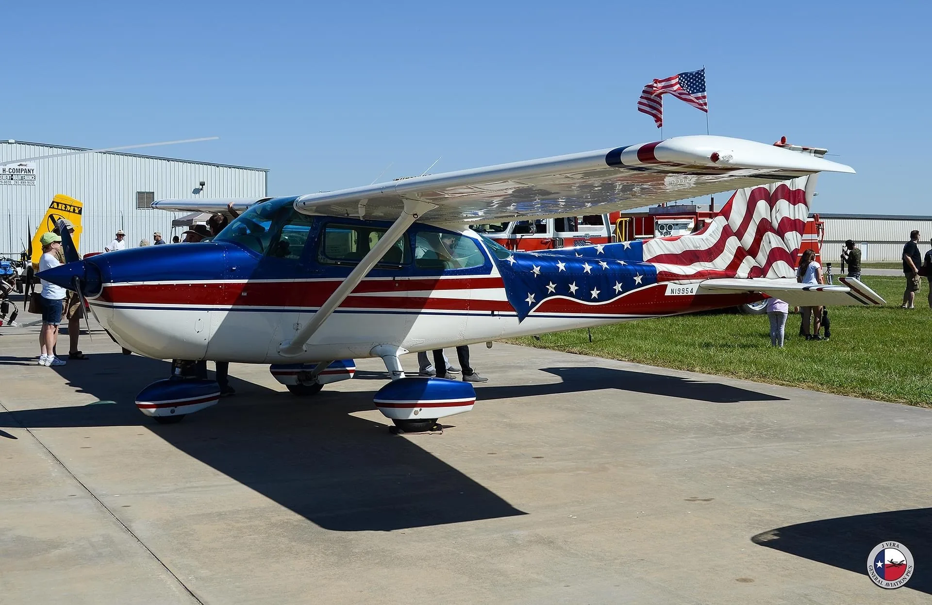 A side view of a Cessna 172 with an American flag paint scheme, parked on a tarmac with people and buildings in the background.