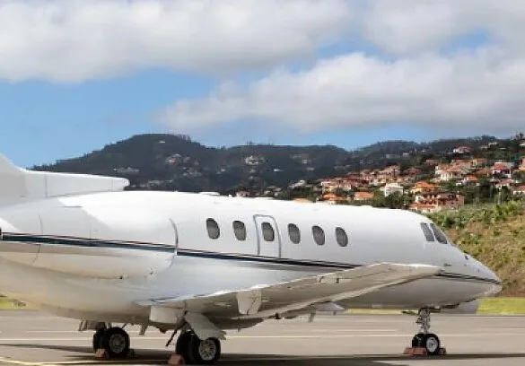 A white business jet with a blue stripe on the tarmac, with a hillside town in the background.