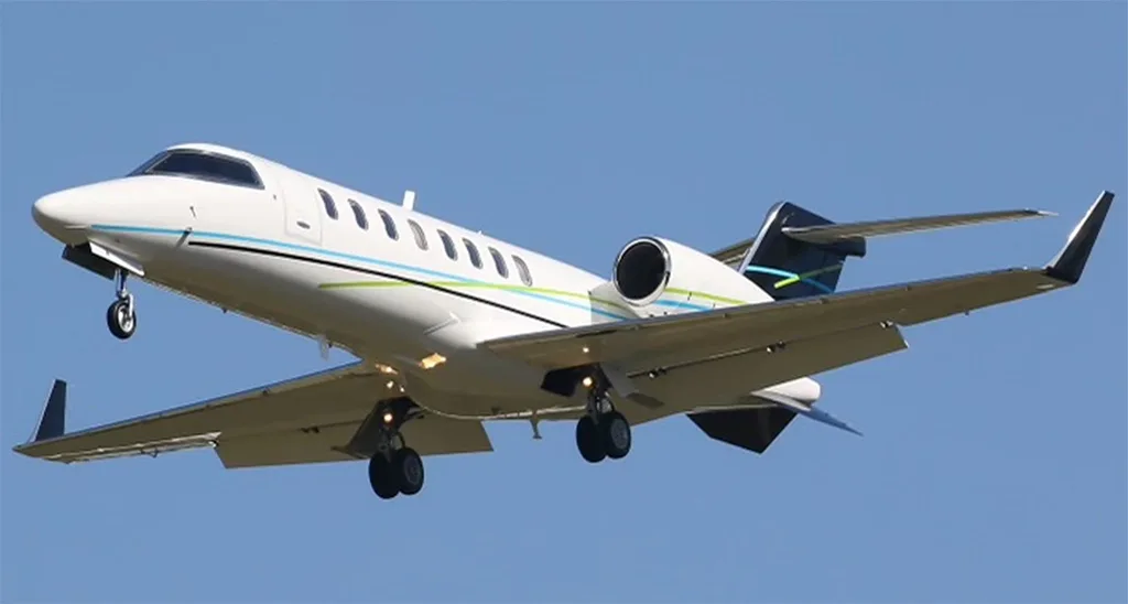A white business jet with blue and green stripes on its fuselage and tail, landing with gear extended against a clear blue sky.