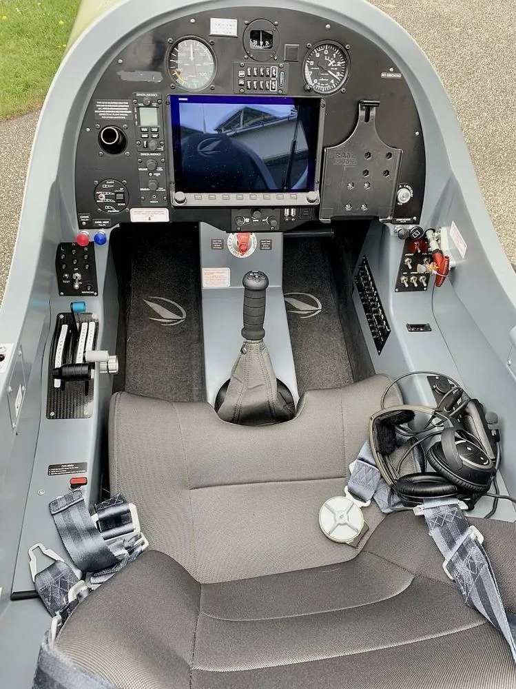 The cockpit of a light aircraft, showing the instrument panel with a central display, flight controls, and a single grey seat with a headset.