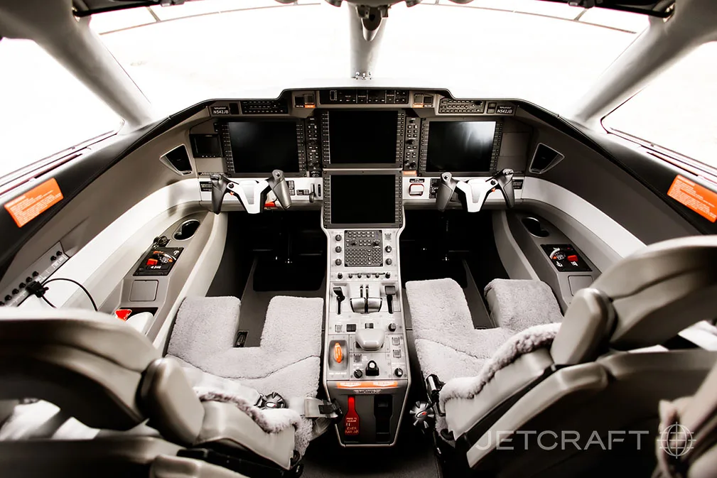 The modern cockpit of a business jet features multiple large display screens and two pilot seats with sheepskin covers.