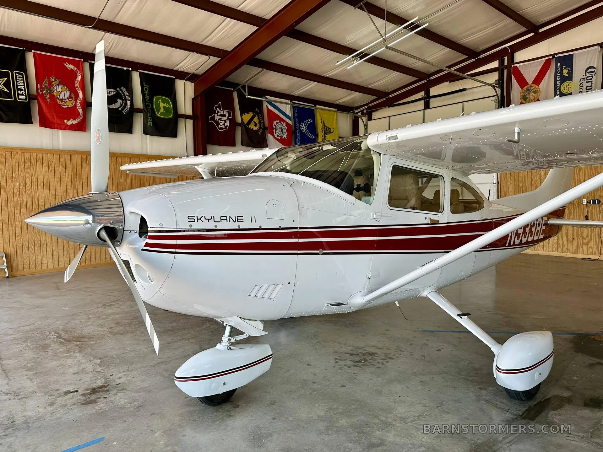 The front-left view of a white Cessna Skylane II with red stripes and a polished propeller spinner, parked in a hangar.