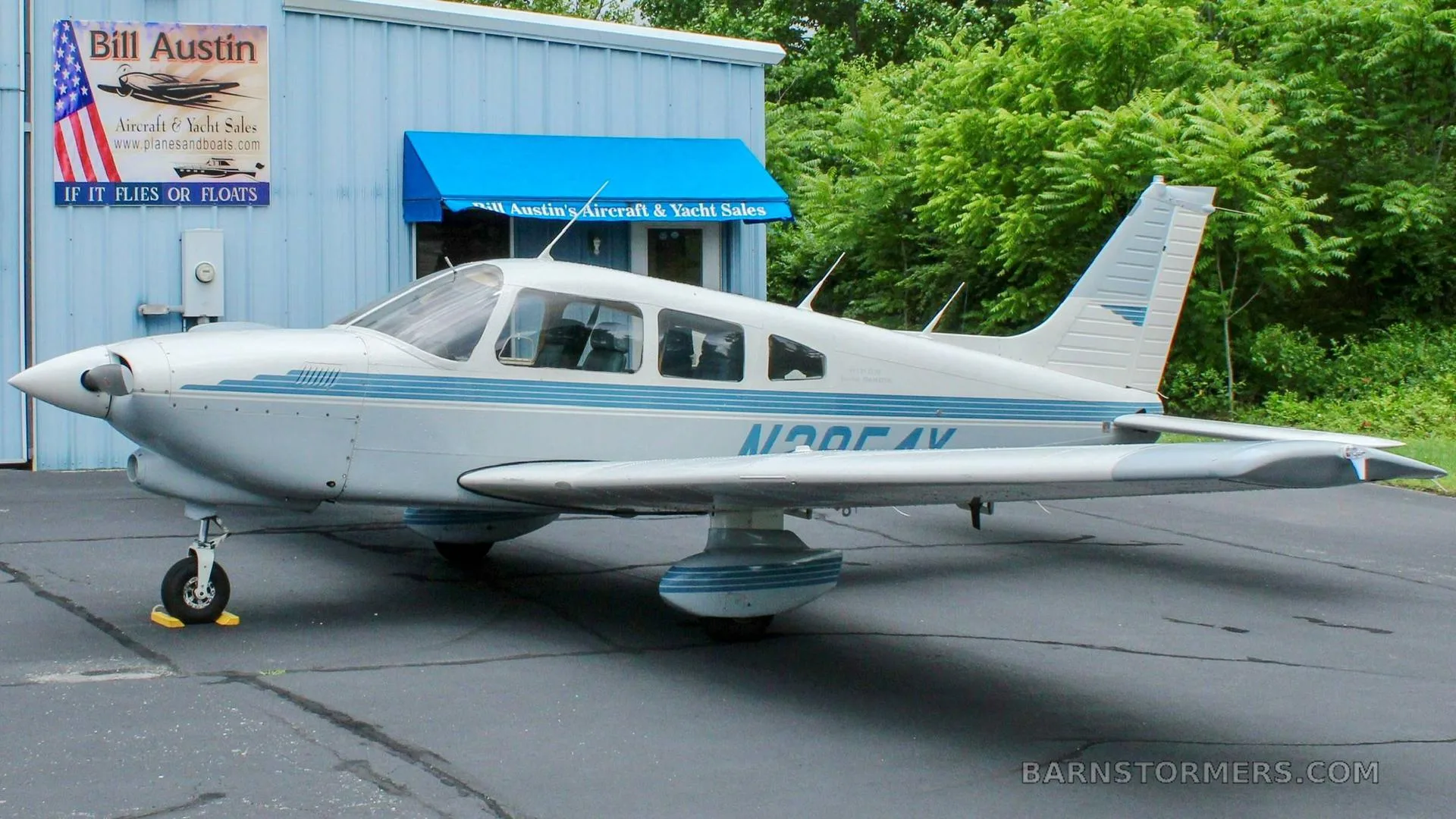 A white single-engine aircraft with blue stripes, registration N2854X, parked on asphalt next to a light blue building.