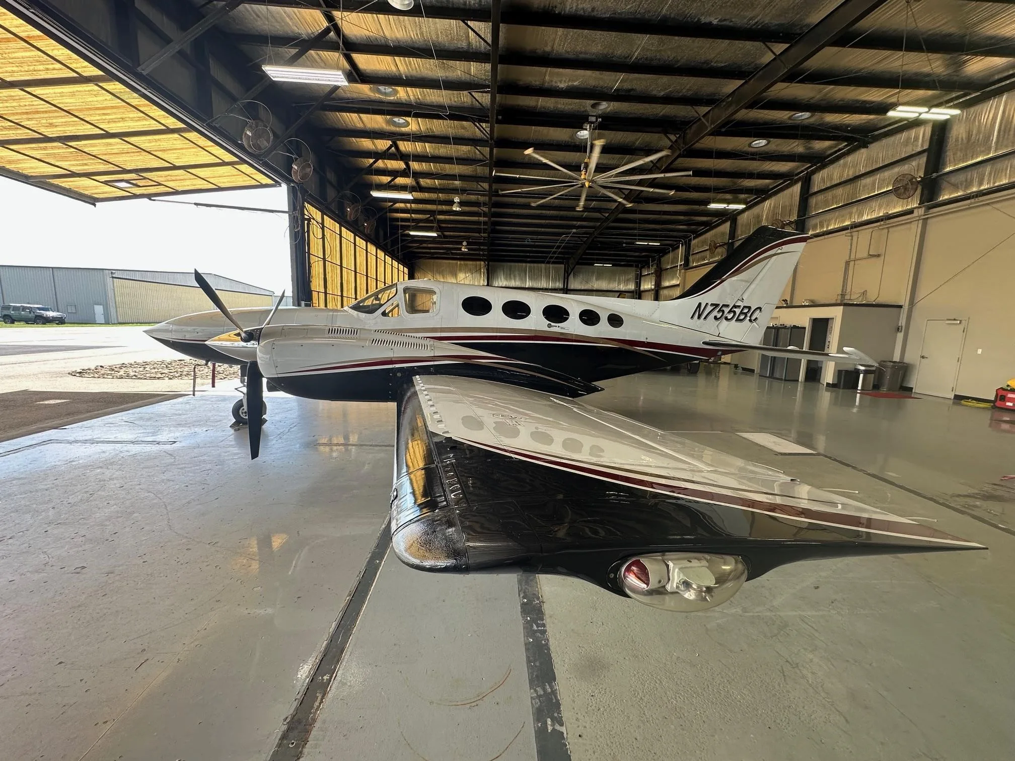 A white and black twin-engine propeller aircraft with registration N7555BC is parked inside a hangar, viewed from the front-right.