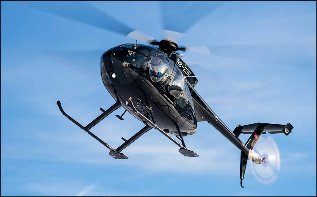 A black Airbus H125 helicopter, registration HB-ZSR, flying against a clear blue sky, viewed from below.