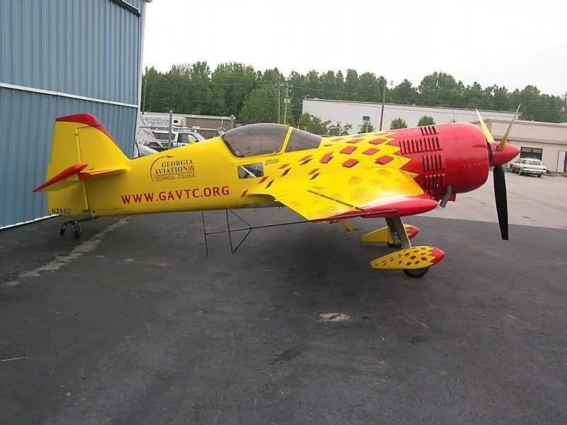 A yellow and red Sukhoi Su-26 aerobatic aircraft with 'GEORGIA AVIATION TECHNICAL COLLEGE' markings, parked on an asphalt tarmac next to a building.