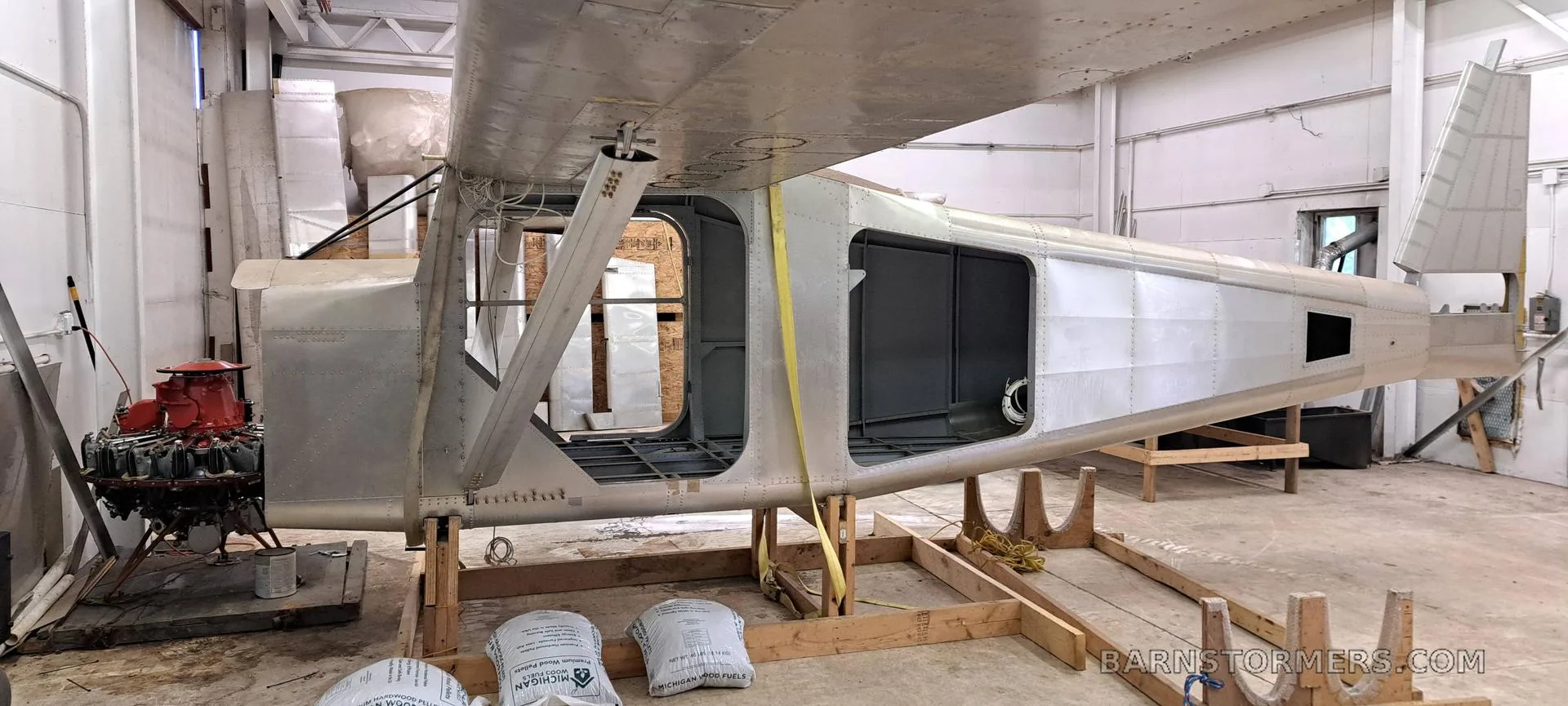 A partially assembled bare metal aircraft fuselage with an exposed red engine on the left, supported on wooden stands in a workshop.