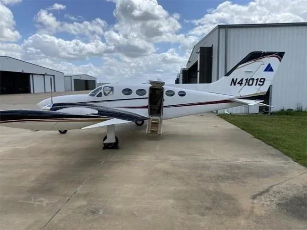 A white twin-engine Cessna 414 Chancellor aircraft with black and gold stripes, parked on an tarmac with its passenger door open, in front of hangars, showing the registration N41019 on the tail.