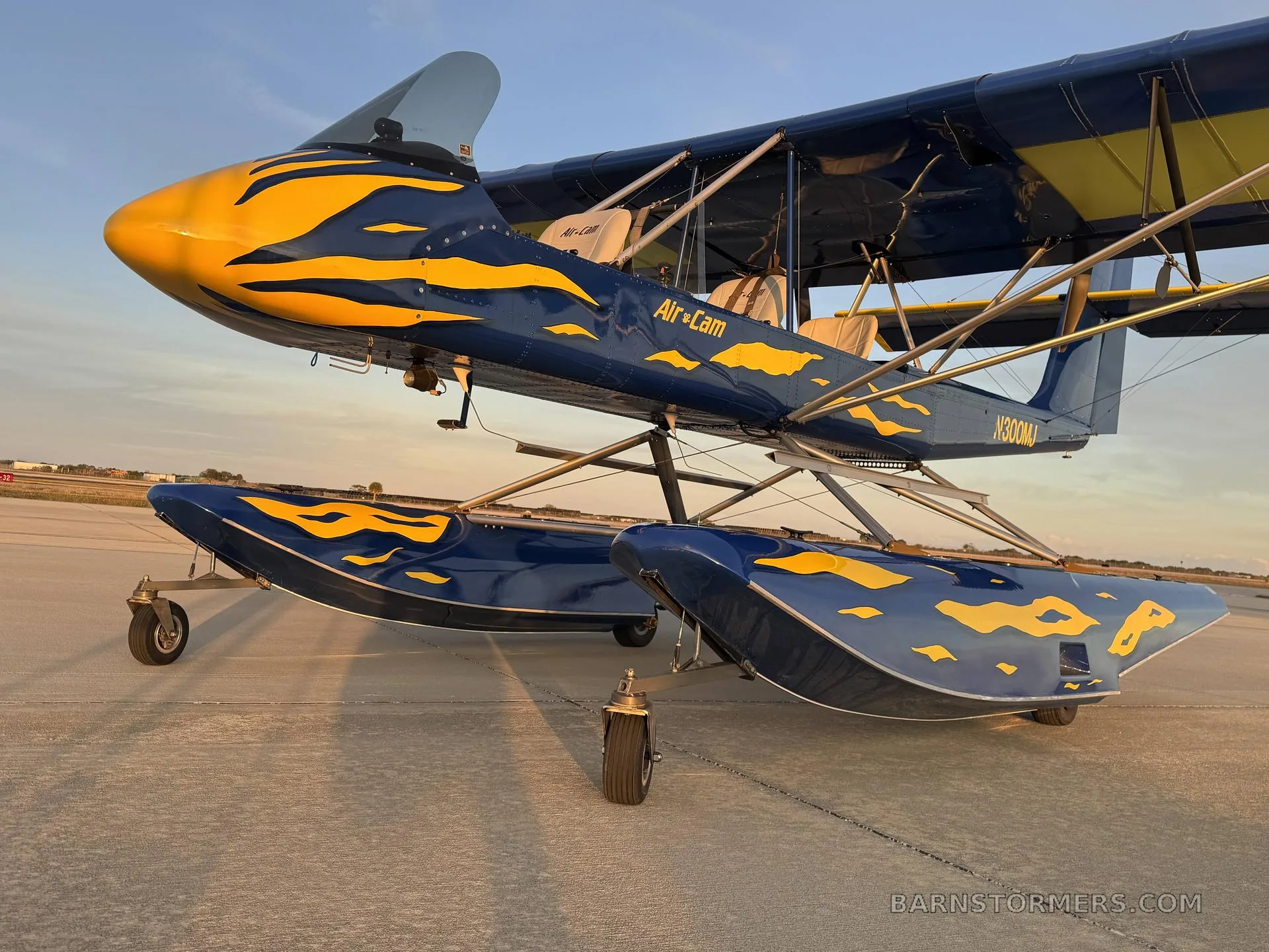 A blue and yellow Air-Cam seaplane with flame decals on floats, viewed from the front-left on an airfield at sunset.