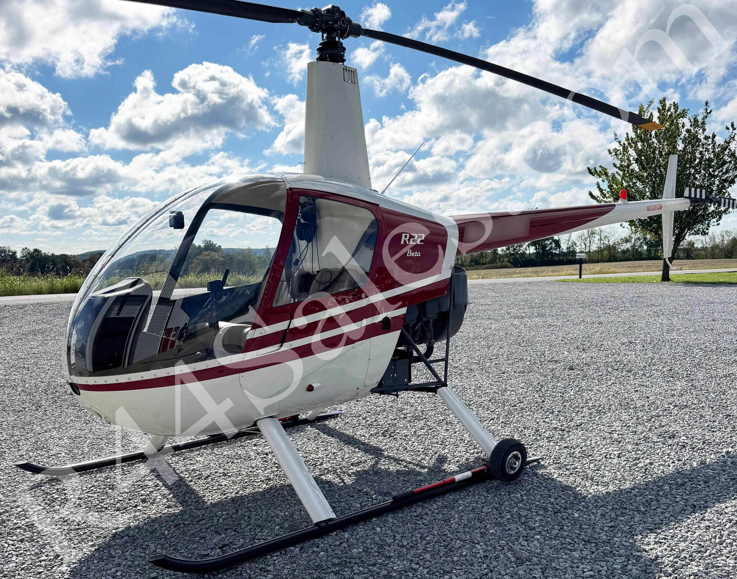 A white and red Robinson R22 Beta helicopter parked on a gravel surface under a partly cloudy sky.