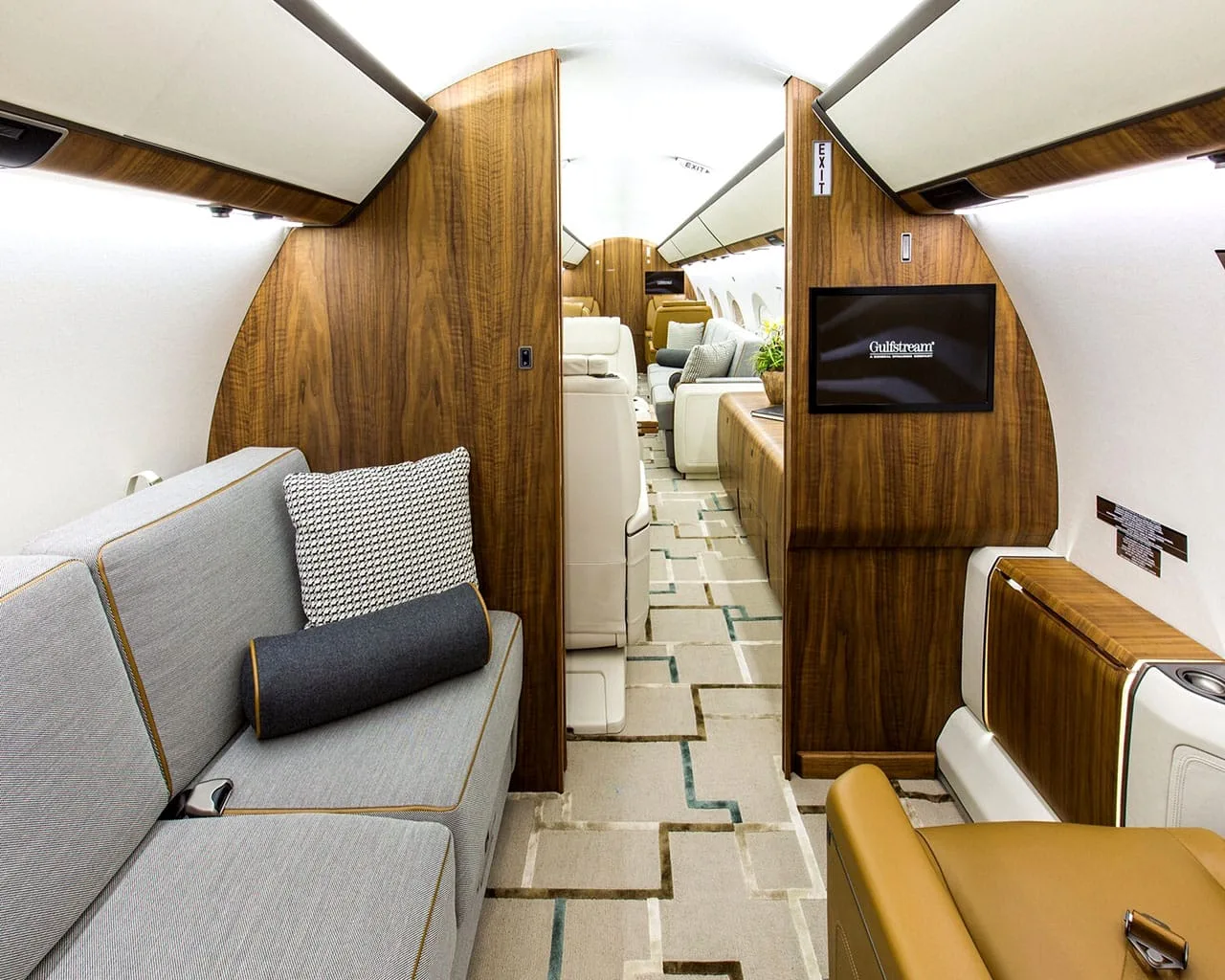 View down the luxurious private jet cabin aisle, showing a grey sofa, brown leather seat, and a screen displaying "Gulfstream".